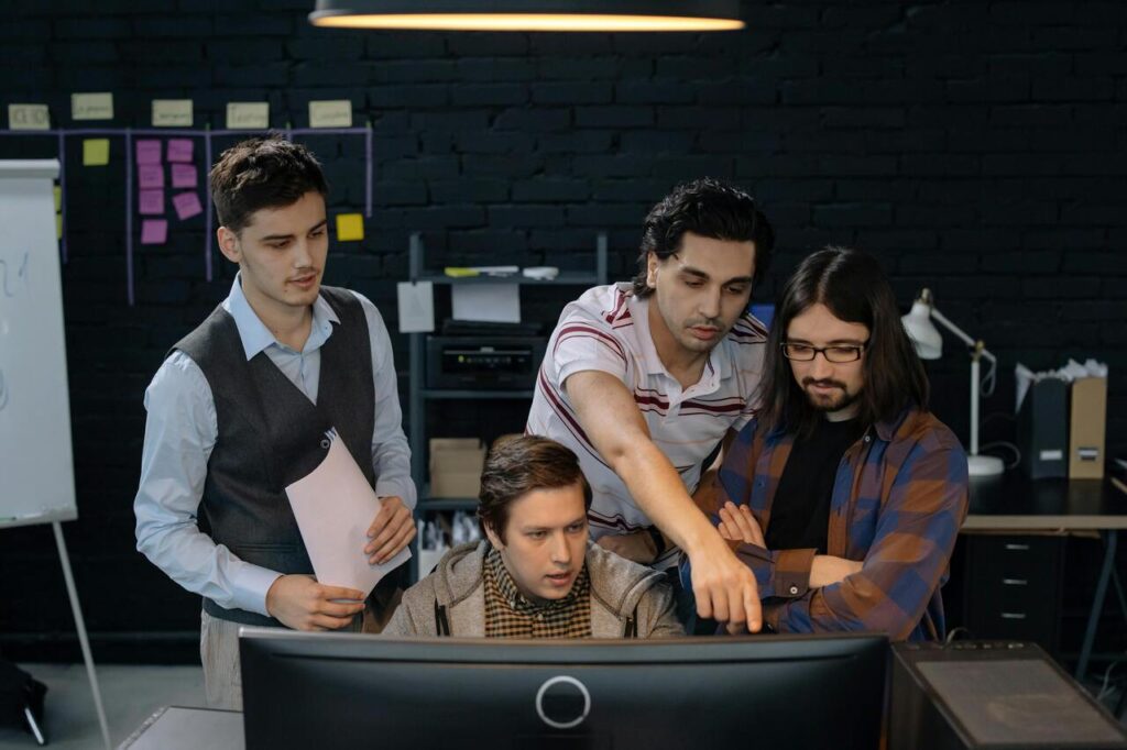 Quatro profissionais colaboram em frente a um monitor em ambiente moderno, com quadro de ideias ao fundo. A cena representa um time engajado em tecnologia ou design, exemplificando o modelo Squad as a Service com foco em soluções ágeis e compartilhadas.